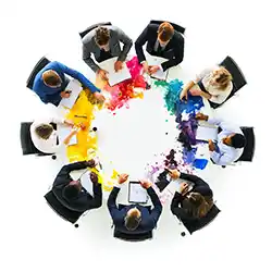 A small business team sits strategizing around a round table with colorful paint spilled around the table.